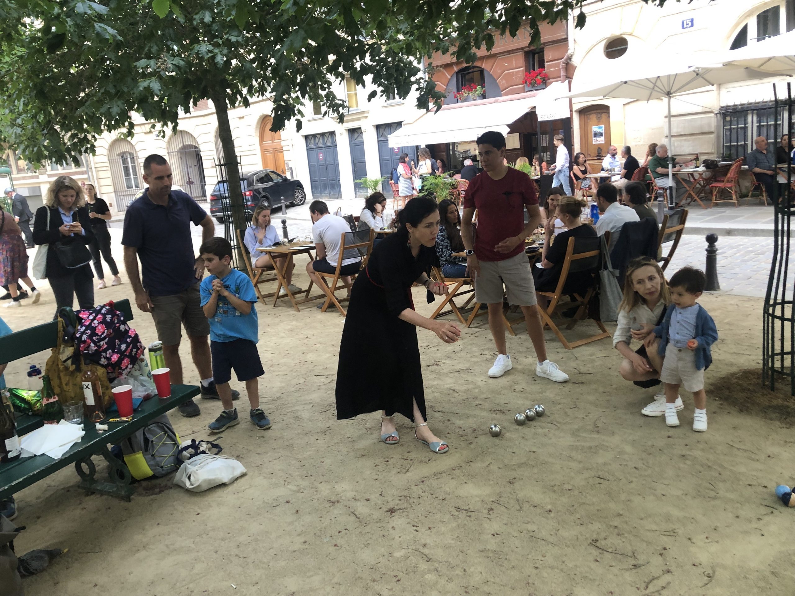 Expat families and children playing pétanque in a kid-friendly square in Paris, surrounded by cafés and locals.